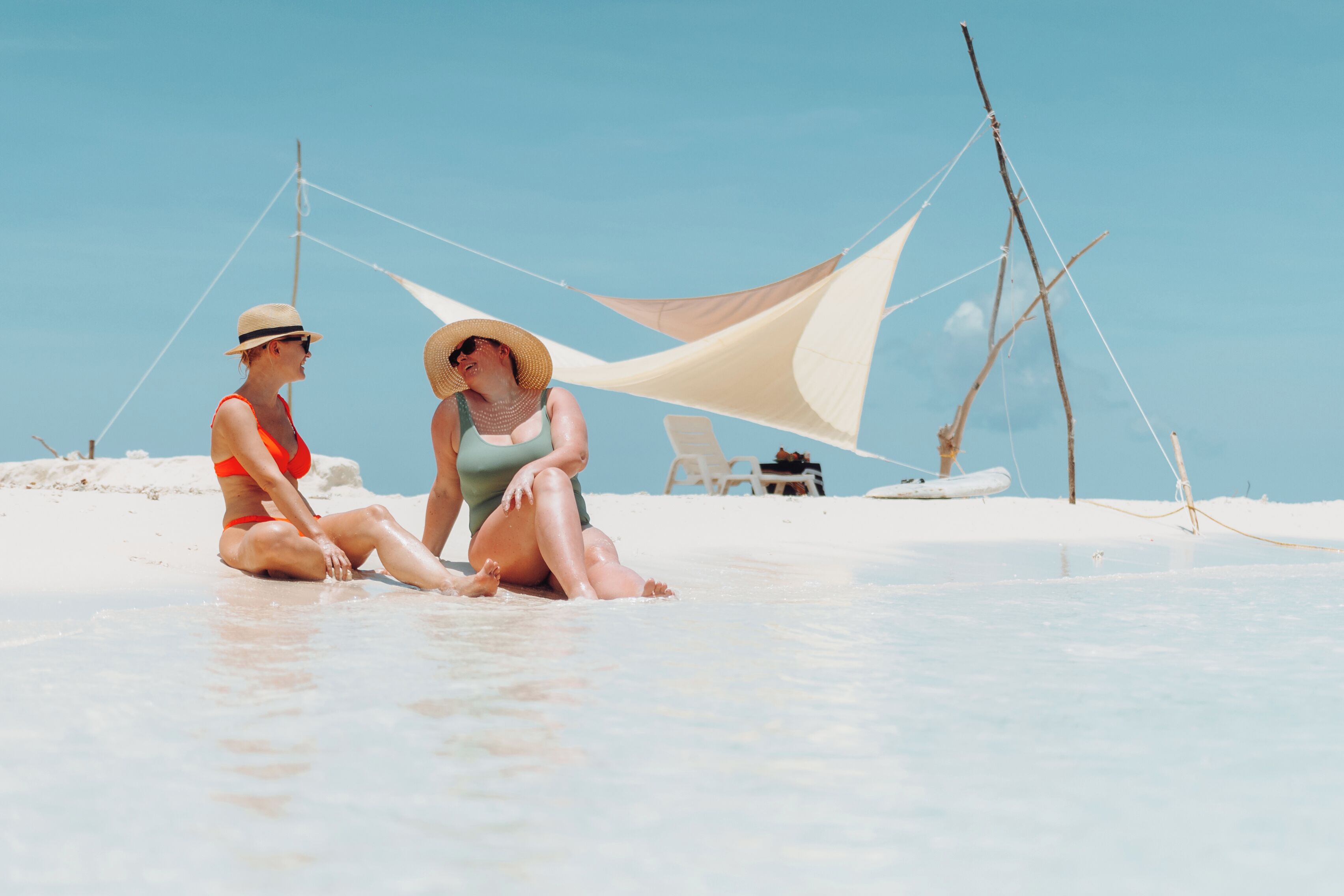 Two people sitting on a beach with a sail shade and a boat in the background.