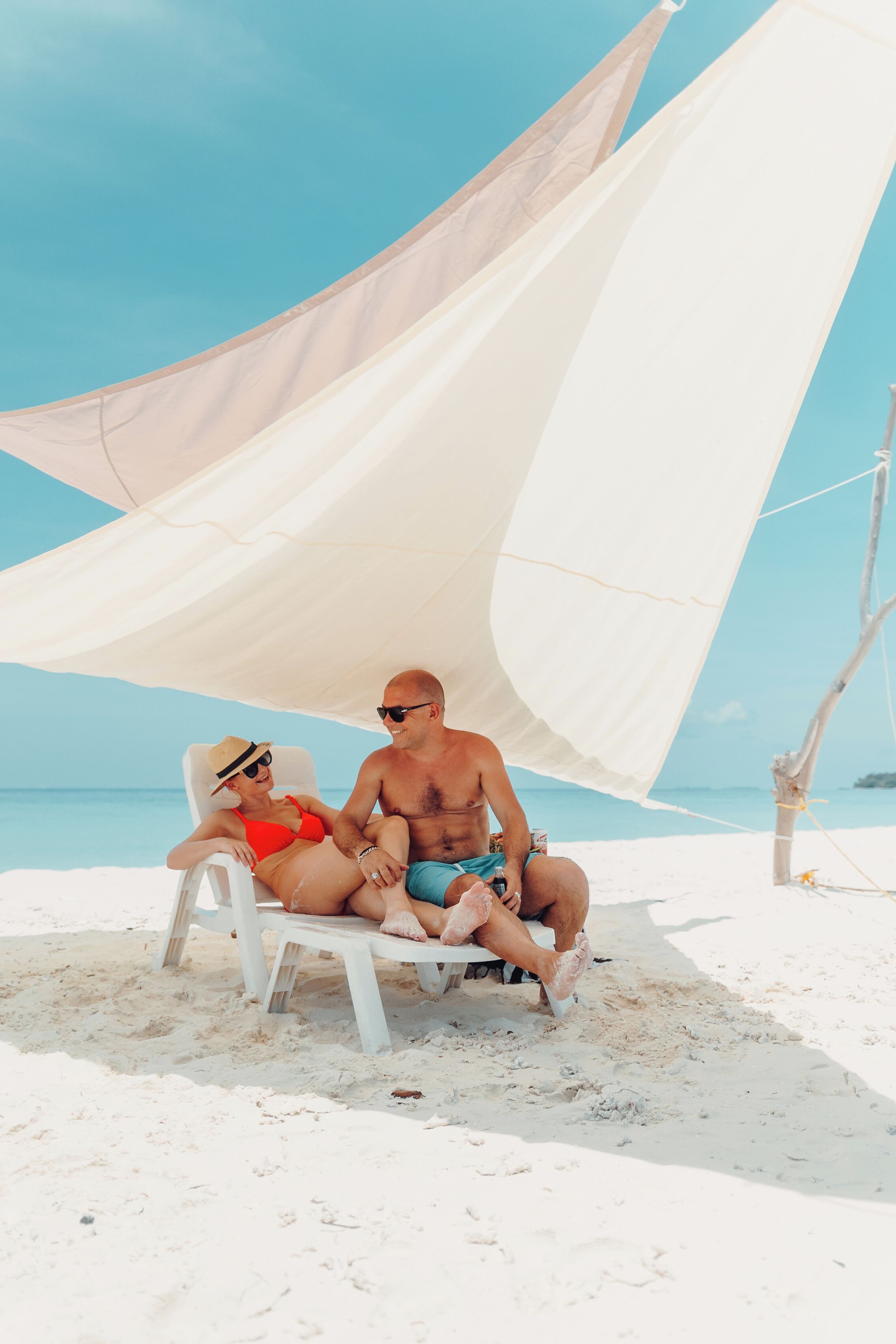 A couple sitting on a white lounge chair under a white canopy on a sandy beach with clear blue water in the background.