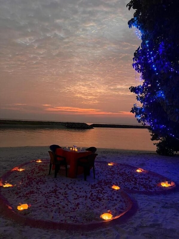 A romantic outdoor dining setup with a table and chairs, surrounded by a circle of candles and rose petals, overlooking a body of water at sunset.