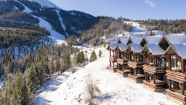A row of wooden cabins with pitched roofs, set against a backdrop of a snow-covered mountain and forest. - Ski-in, Ski-out Townhome with Private Hot Tub (Big Sky)