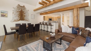 A modern living room with a dining area, wooden beams, a large rug, and a cowhide wall art piece. - Ski-in, Ski-out Townhome with Private Hot Tub (Big Sky)
