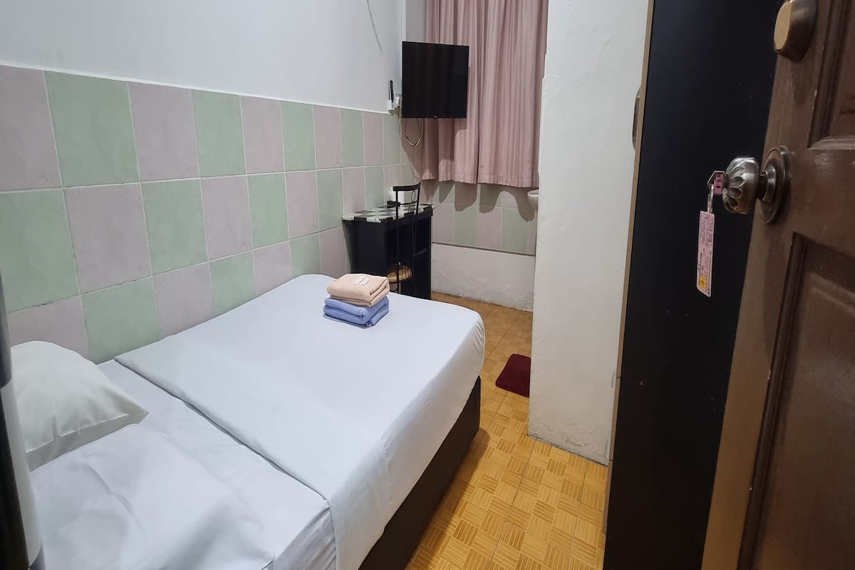 Ein kleiner, kompakter Raum mit einem Bett, einem Sessel, einem kleinen Tisch und einem an der Wand befestigten Fernseher.