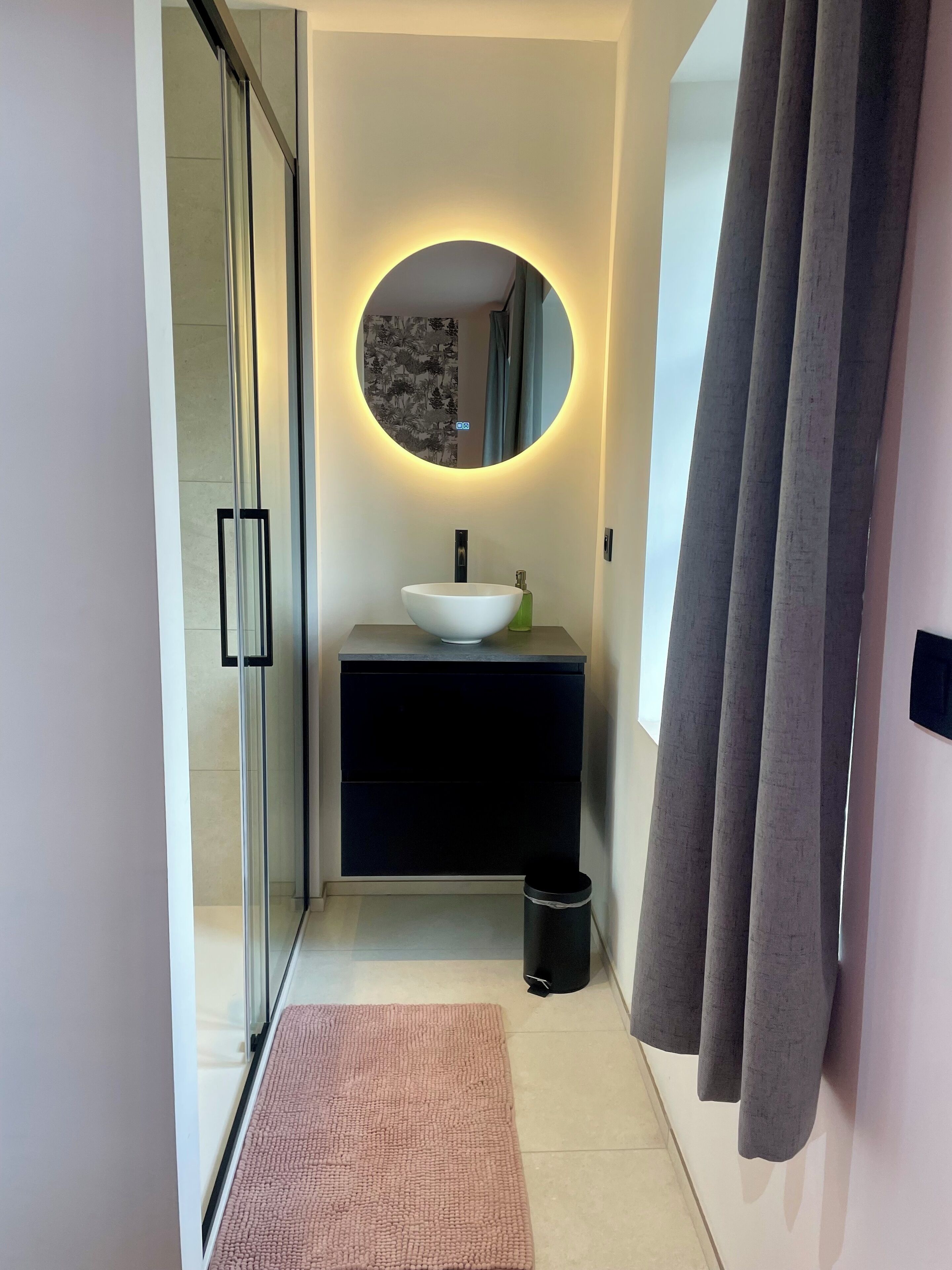 Une salle de bain moderne avec une douche à parois de verre, un grand miroir rond doté d’un éclairage indirect, un lavabo blanc et un meuble-lavabo noir.