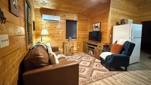 A cozy cabin interior with a brown leather sofa, a blue armchair, a wooden entertainment unit, and a white refrigerator.