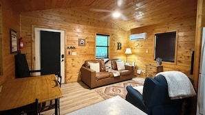 A cozy cabin interior with a wooden ceiling, a brown leather sofa, a dining table, and a blue armchair.