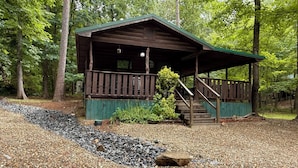 A wooden cabin with a porch and stairs, surrounded by trees and pebbles.