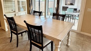 A dining area with a large table, four chairs, a hanging light fixture, and a view of the outdoors through glass doors.