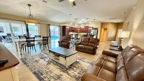 A spacious living room with a brown leather sofa, a coffee table, and a dining area with chairs and a table. There is a kitchen in the background with cabinets and a ceiling fan.