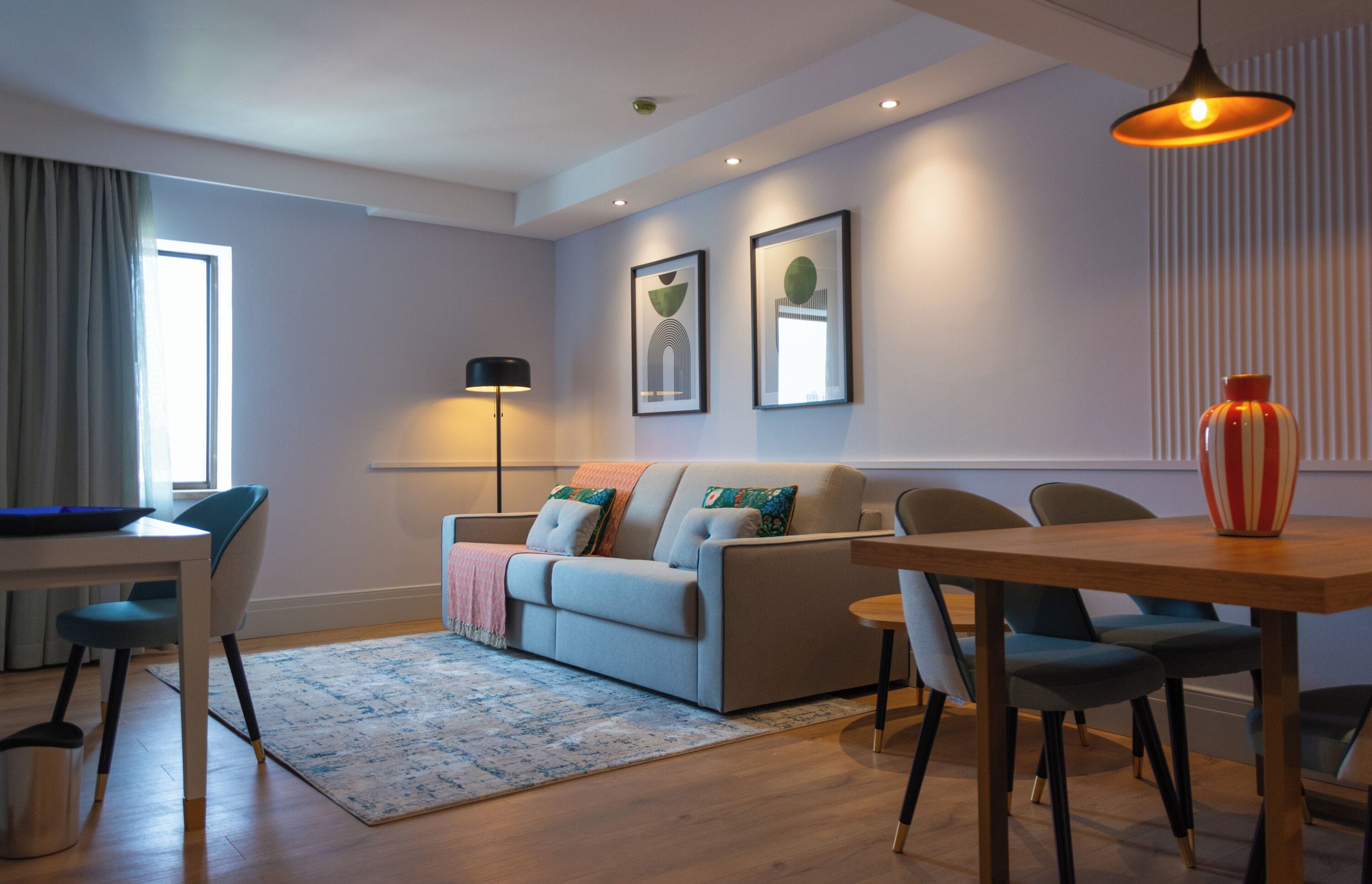 A modern living room with a sofa, coffee table, and chairs. There is a window with curtains, a floor lamp, and framed artwork on the wall.