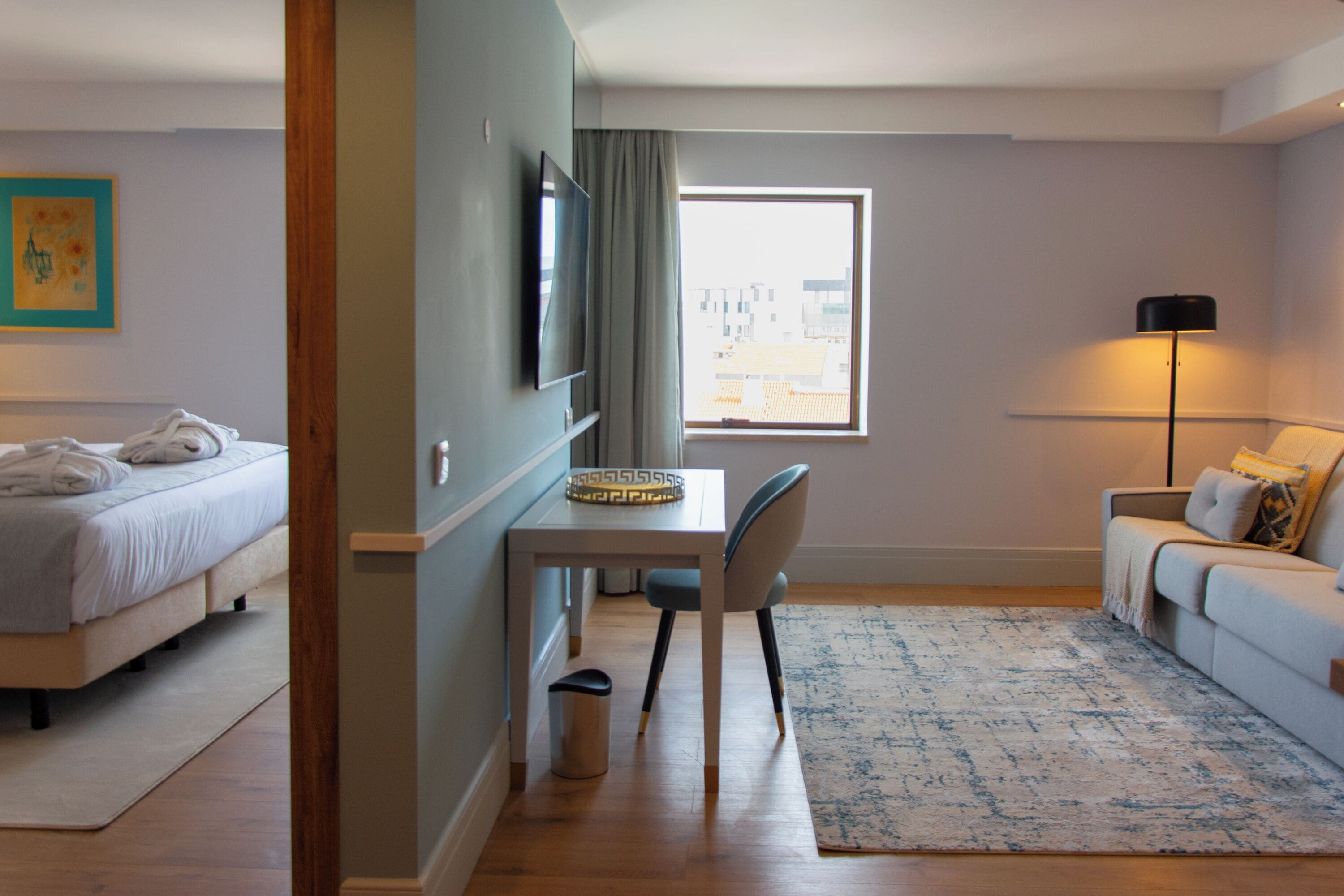 Habitación de hotel con cama, un escritorio con silla, televisión, lámpara y ventana con vistas a edificios.