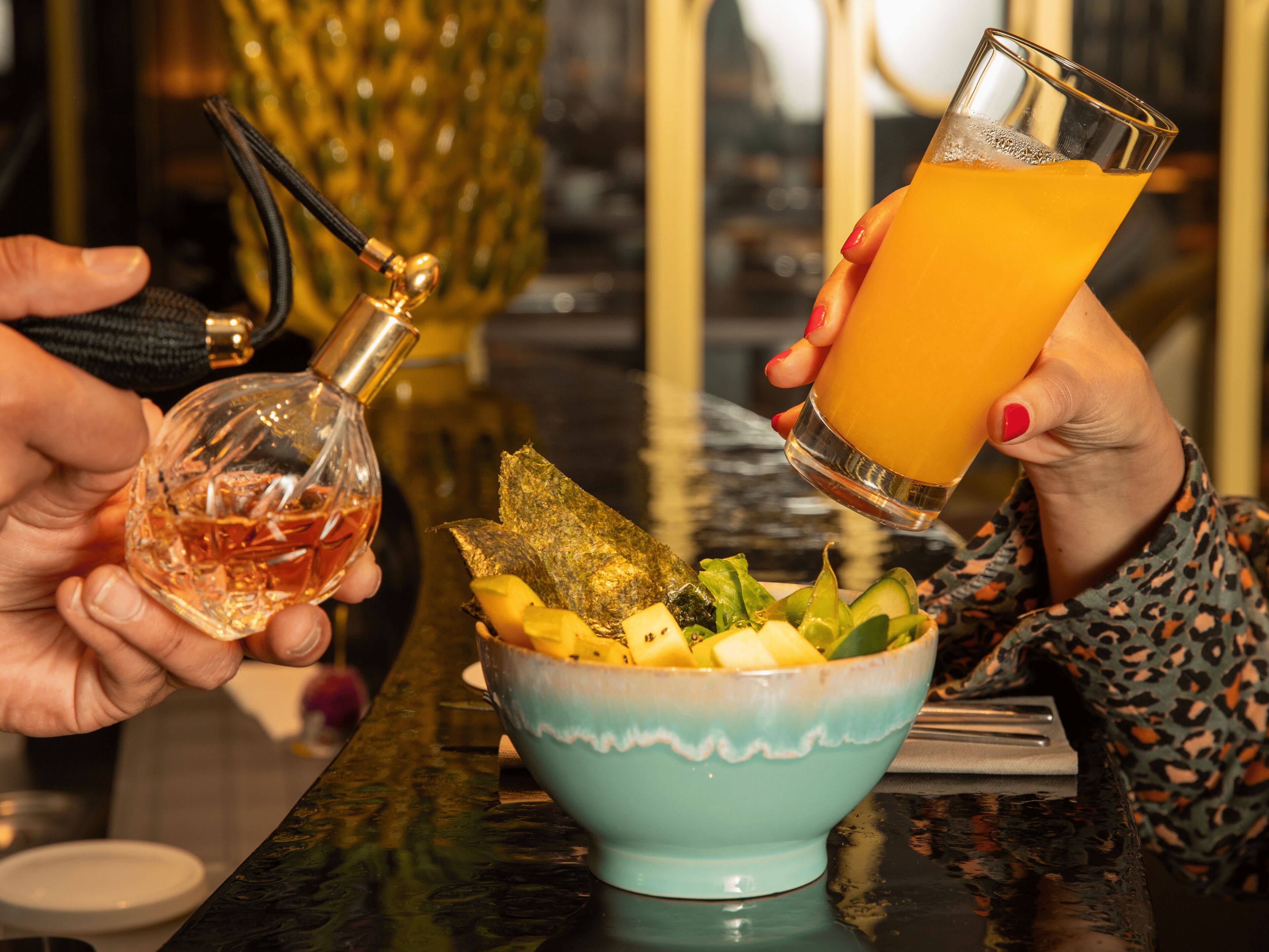 En person hælder drik i et glas, holder en flaske spiritus, og en skål med frugt står på et bord.