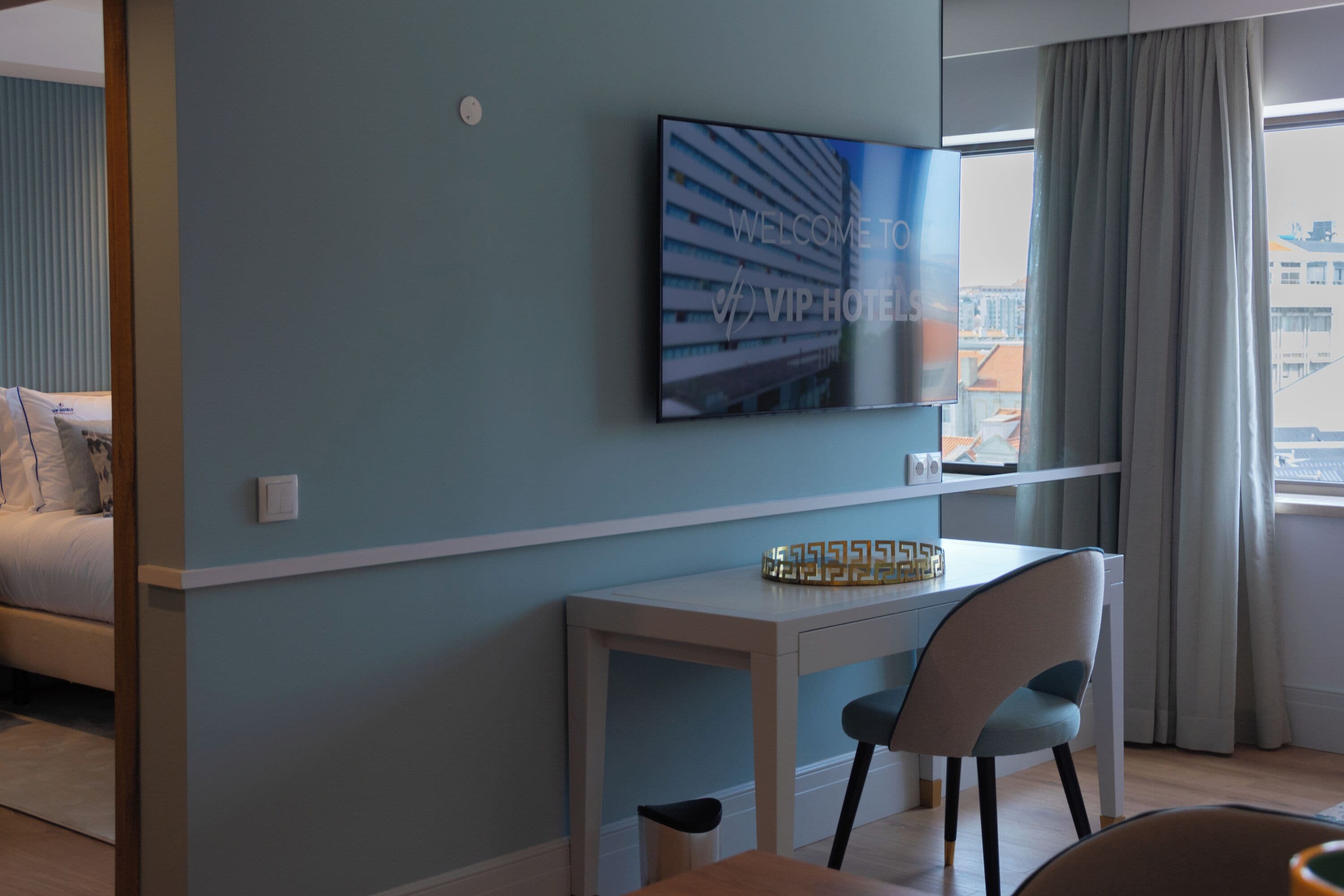 A modern hotel room with a bed, a desk with a chair, a TV mounted on the wall, and a window with curtains.