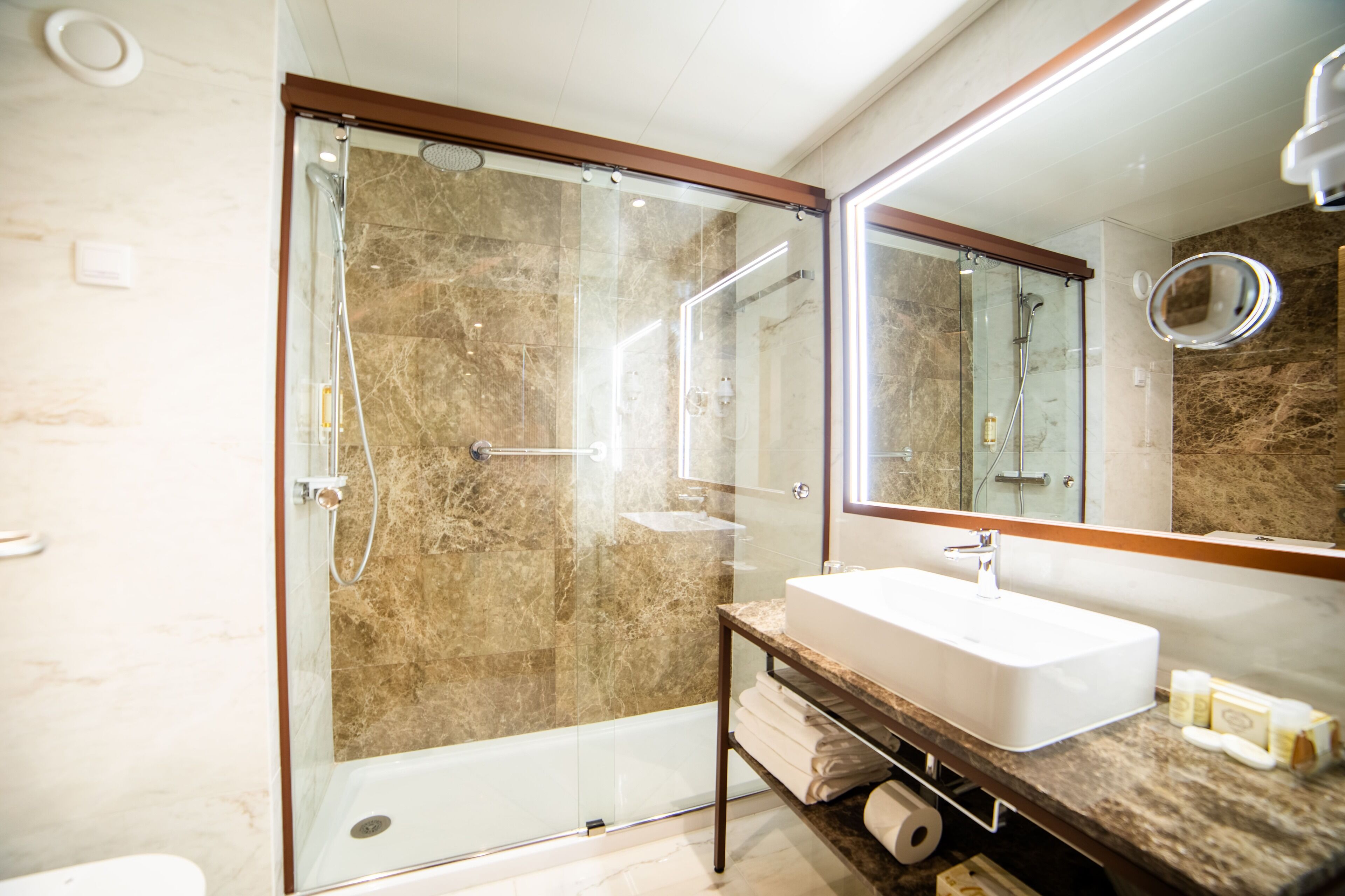 A modern bathroom with a glass-enclosed shower, a marble-tiled wall, a white sink, and a large mirror.