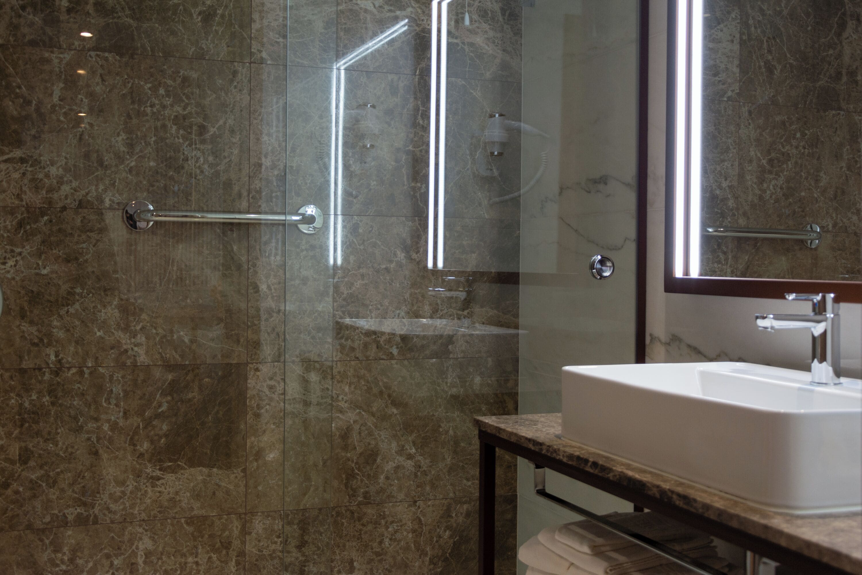 Une salle de bain moderne avec une douche à parois de verre, un mur recouvert de carreaux de marbre, un lavabo blanc et un grand miroir.