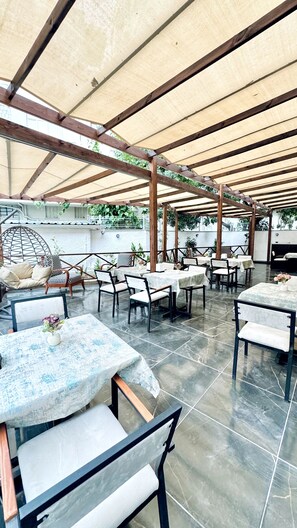 Een terras buiten met tafels en stoelen onder een houten pergola met een beige dakbedekking.