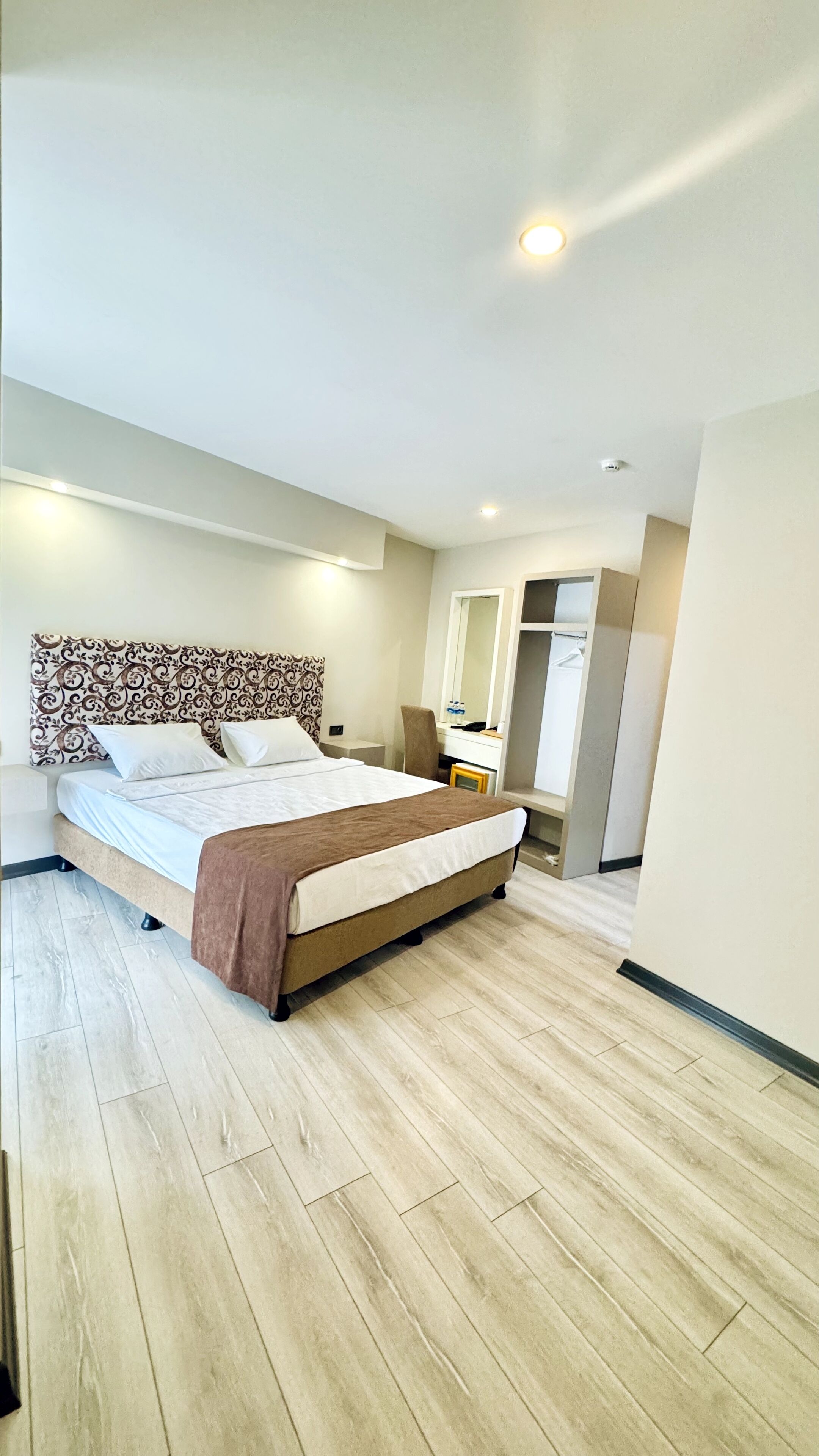 A modern hotel room with a large bed, a desk, a chair, a mirror, and a wardrobe.