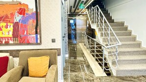 Um hall de hotel com piso de mármore, um sofá com almofada amarela, escada com corrimão de metal e uma pintura abstrata colorida na parede.