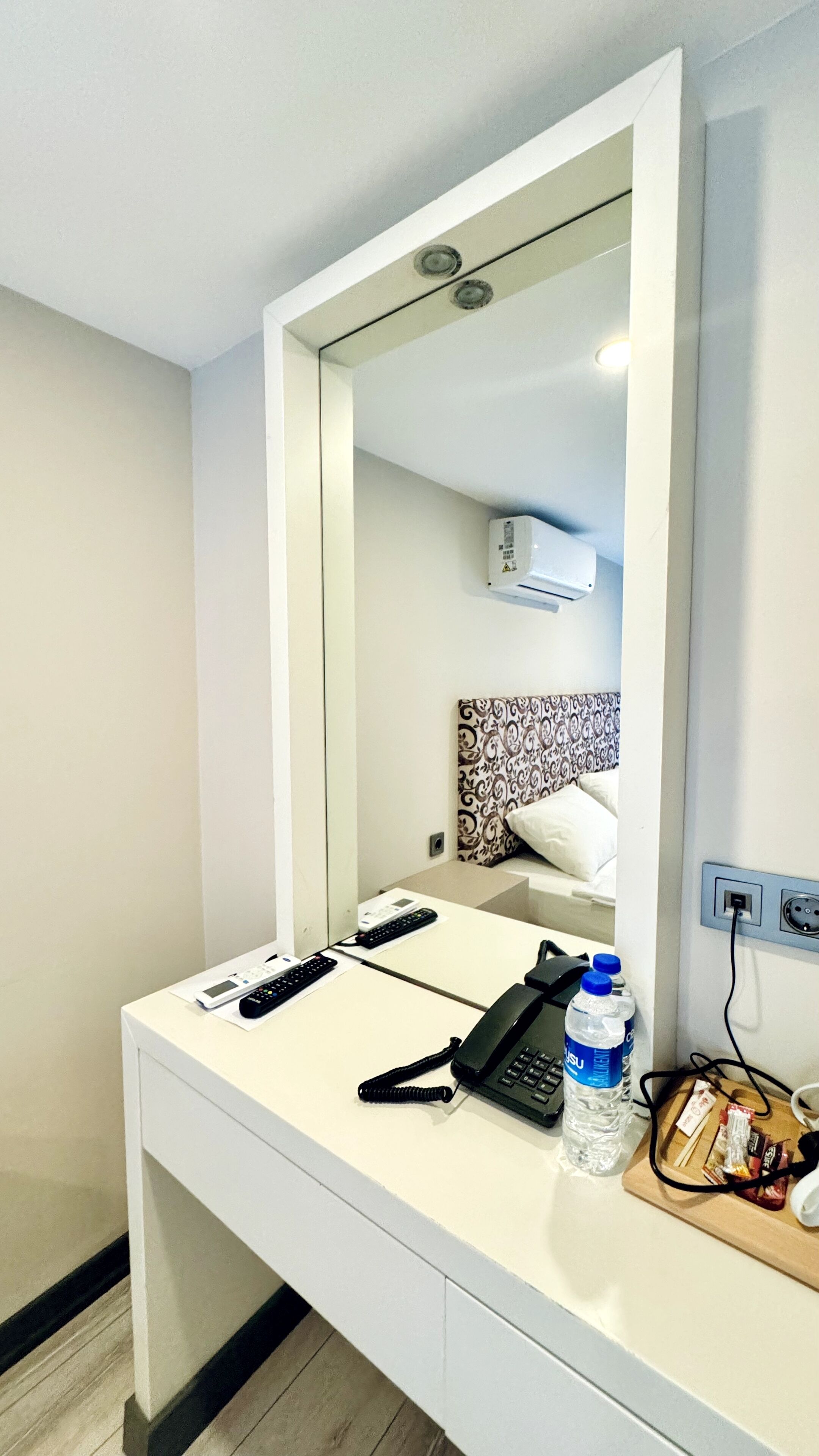 Habitación de hotel con un escritorio blanco, teléfono, control remoto, botella de agua y un espejo que refleja la habitación.