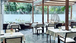 Restaurante ao ar livre com mesas e cadeiras sob uma pérgola sombreada, uma cadeira suspensa e vista para um corpo d'água.