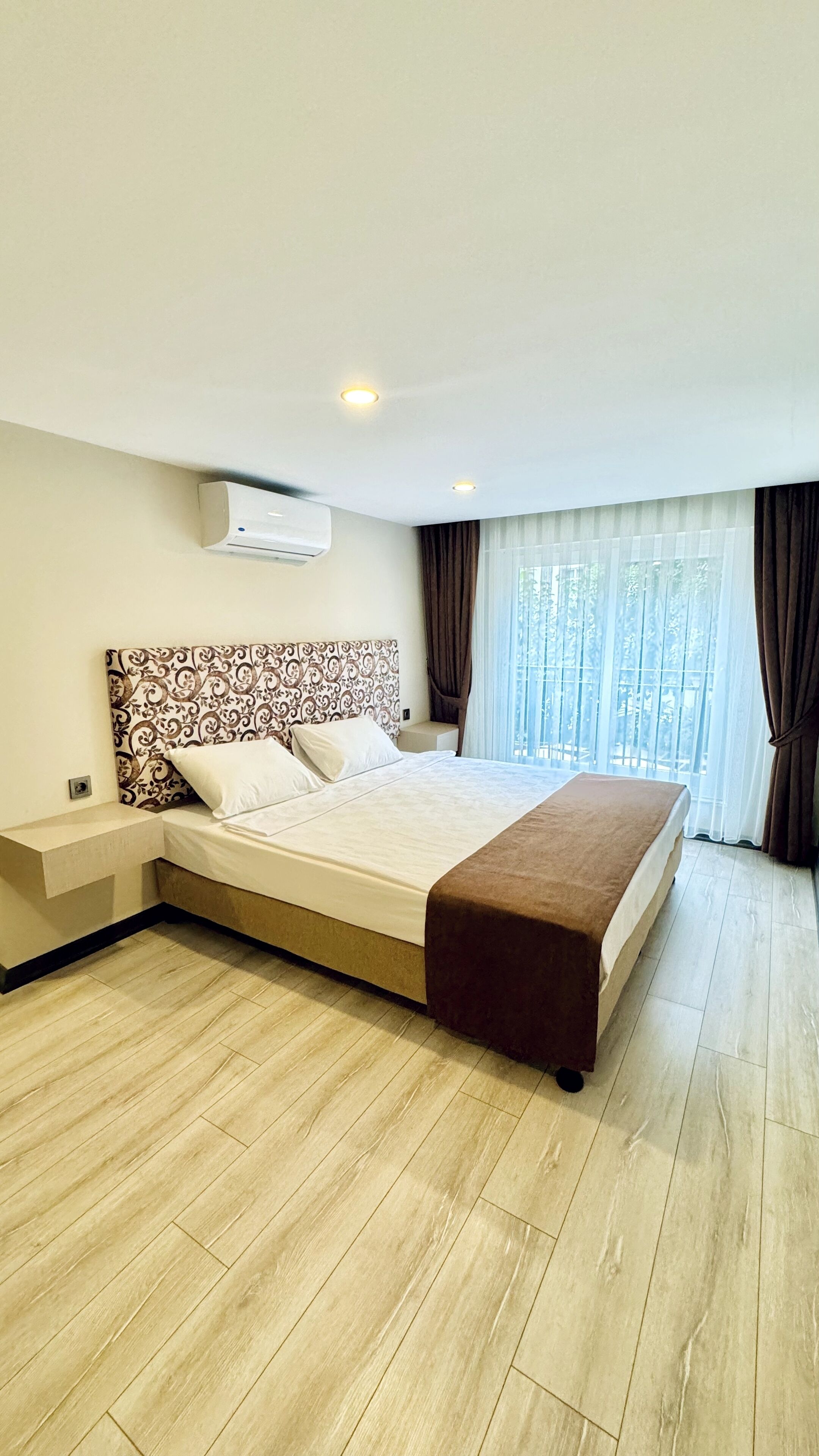 A modern hotel room with a large bed, a bedside table, an air conditioning unit, and a window with curtains.
