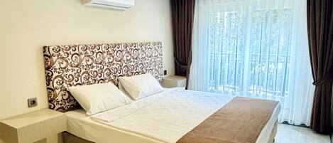 A modern hotel room with a large bed, a bedside table, an air conditioning unit, and a window with curtains.