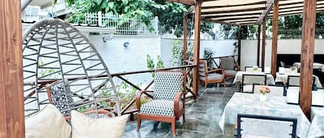 Un patio al aire libre con pérgola de madera, asientos acolchados y suelo de baldosas.