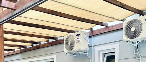 Um pátio externo com mesas e cadeiras, coberto por uma pérgola de madeira e equipado com ventiladores de teto.