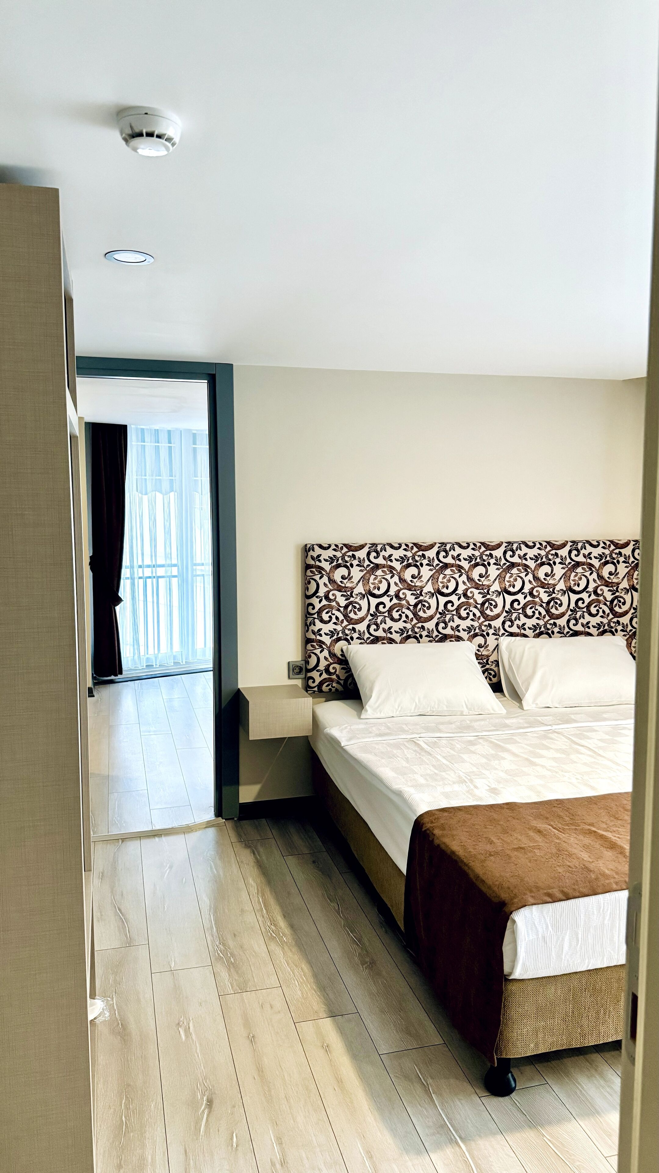 A hotel room with a large bed, a headboard with a patterned design, a wooden floor, and a view of the outside through a glass door.