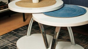 Een moderne woonkamer met een donkerblauwe bank, een witte vaas met gedroogde bloemen en twee ronde tafels met geweven onderzetters.