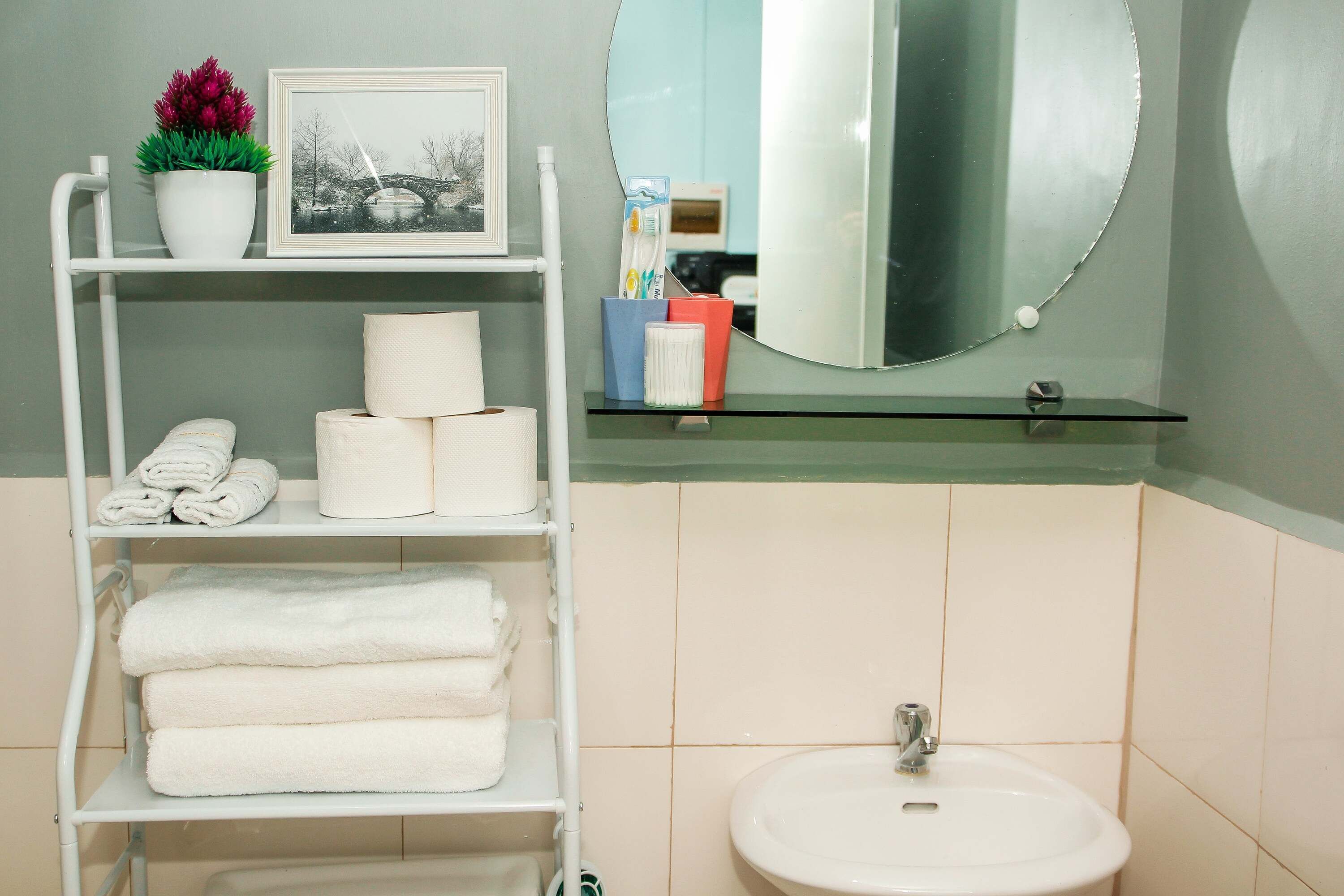 Une salle de bain avec une étagère blanche où sont posées des serviettes, un tableau encadré et une plante en pot.