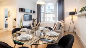 A modern living room with a glass dining table set for four, a sofa, a TV mounted on the wall, and a large window with curtains. - Premier Three Bedroom Bliss City View Shoreditch (London)