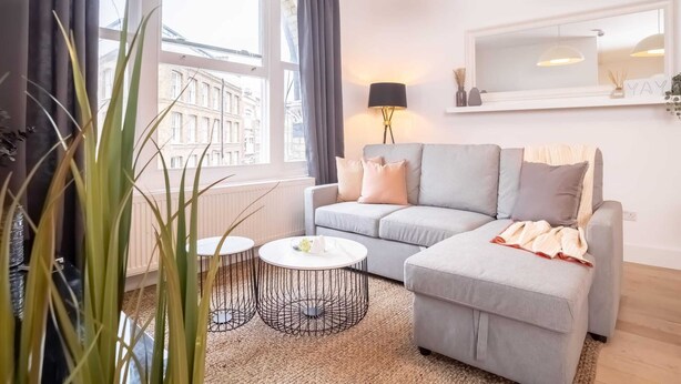 A modern living room with a grey sofa, two round coffee tables, and a large window with curtains. - Premier Three Bedroom Bliss City View Shoreditch (London)