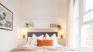 A neatly made bed with white and orange pillows, a headboard with a shelf above, and a window with a view of buildings. - Premier Three Bedroom Bliss City View Shoreditch (London)