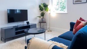 Ein modernes Wohnzimmer mit Flachbildfernseher, einer dunkelgrauen Unterhaltungseinheit, einem runden Couchtisch, einem blauen Sofa mit bunten Kissen, einer Topfpflanze und einer Stehlampe.