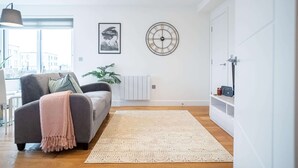 A modern living room with a grey sofa, a large wall clock, and a view of the city through large windows. - One Bed - Queensmere Shopping Centre by Belvilla (Slough)