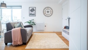 A modern living room with a grey sofa, a large window, a wall clock, and a plant. - One Bed Near Windsor Castle by Belvilla (Slough)