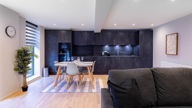 Uma sala de estar moderna com um sofá cinza, uma mesa de jantar posta com pratos e copos, uma cozinha com armários escuros e um vaso com planta.
