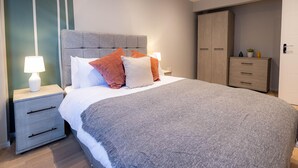 Quarto moderno com uma cama grande, cabeceira cinza, mesas de cabeceira com luminárias, um guarda-roupa e uma pequena planta no aparador.