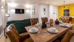 A dining area with a wooden table set for four, surrounded by leather chairs. A green sofa is against the wall, and a flat-screen TV is mounted above it. - Three Bedroom Near Brick Lane by Belvilla (London)