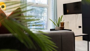 A modern living room with a large window, a flat-screen TV mounted on the wall, and a potted plant on a side table. - Discover Angel Central Two Bedroom by Belvilla (London)