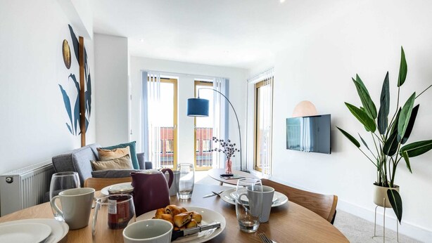 Un comedor con una mesa redonda para dos personas, un sofá al fondo y un ventanal con vistas a edificios.