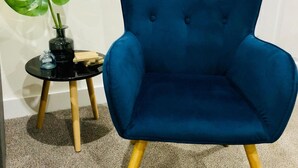 A blue upholstered armchair with wooden legs, positioned next to a small black table with a green plant and a glass vase.