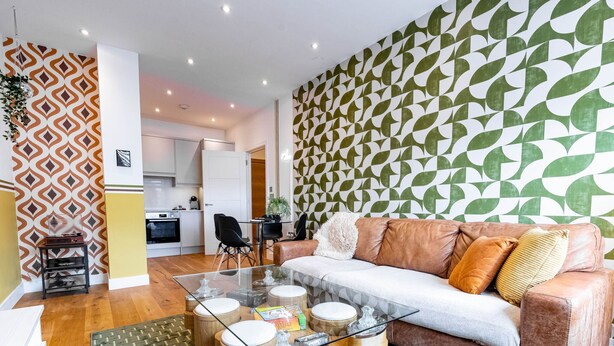 Una sala de estar moderna con una mesa de centro de vidrio, un sofá de cuero, papel tapiz estampado y una cocina al fondo.