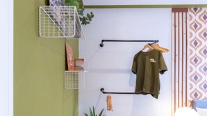 Una habitación con pared verde, puerta blanca, planta colgante, estante blanco con planta, mesa blanca con lámpara y cama con cortina estampada.
