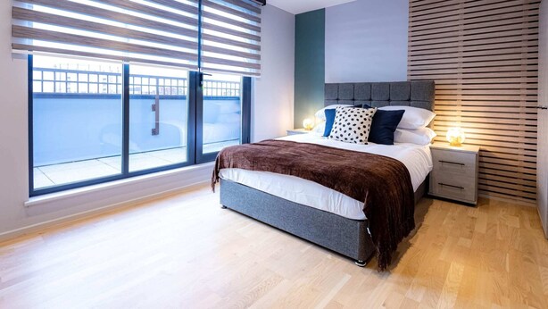 A modern bedroom with a large window, a bed with a grey headboard, and a bedside table with a lamp. - Victoria Park Vista One-bedroom by Sojo Stay (London)