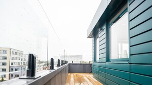 Un balcón con piso de madera, baranda metálica y vista a edificios al fondo.