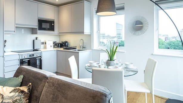 Una cocina moderna con armarios blancos, microondas y un comedor con mesa y sillas de vidrio.