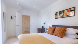 A bedroom with a large bed, a nightstand, a painting, and a door leading to another room. - Horniman Museum Harmony Two-bedroom by Sojo Stay (Mitcham)