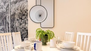 Une table à manger est dressée avec de l’assiette blanche, des bols, des verres et des couverts. On y voit deux chaises, une fougère en pot et un miroir fixé au mur.