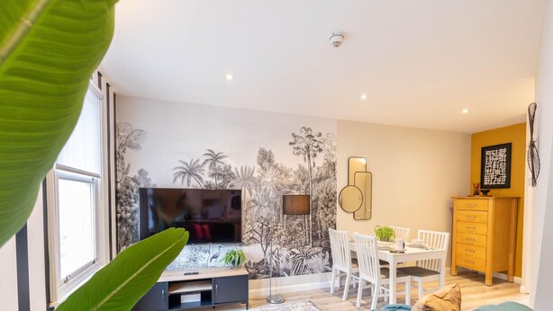 Un salon moderne doté d’un téléviseur à écran plat, d’un coin repas avec une table et des chaises blanches, et d’une cuisine avec des meubles en bois.